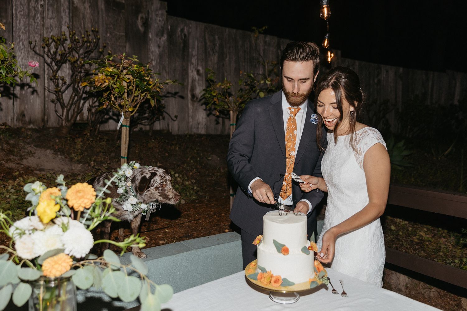 A couple slices into their tiered white wedding cake decorated with flowers at a nighttime outdoor celebration.