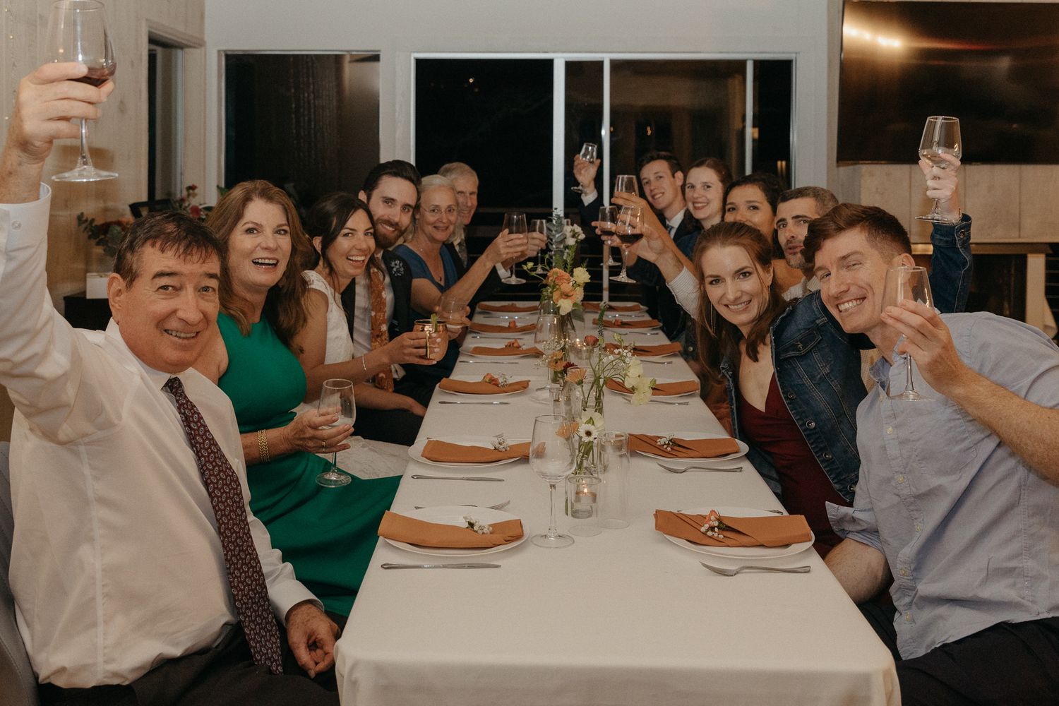 Wedding guests raise their glasses in celebration at a long reception dinner table.