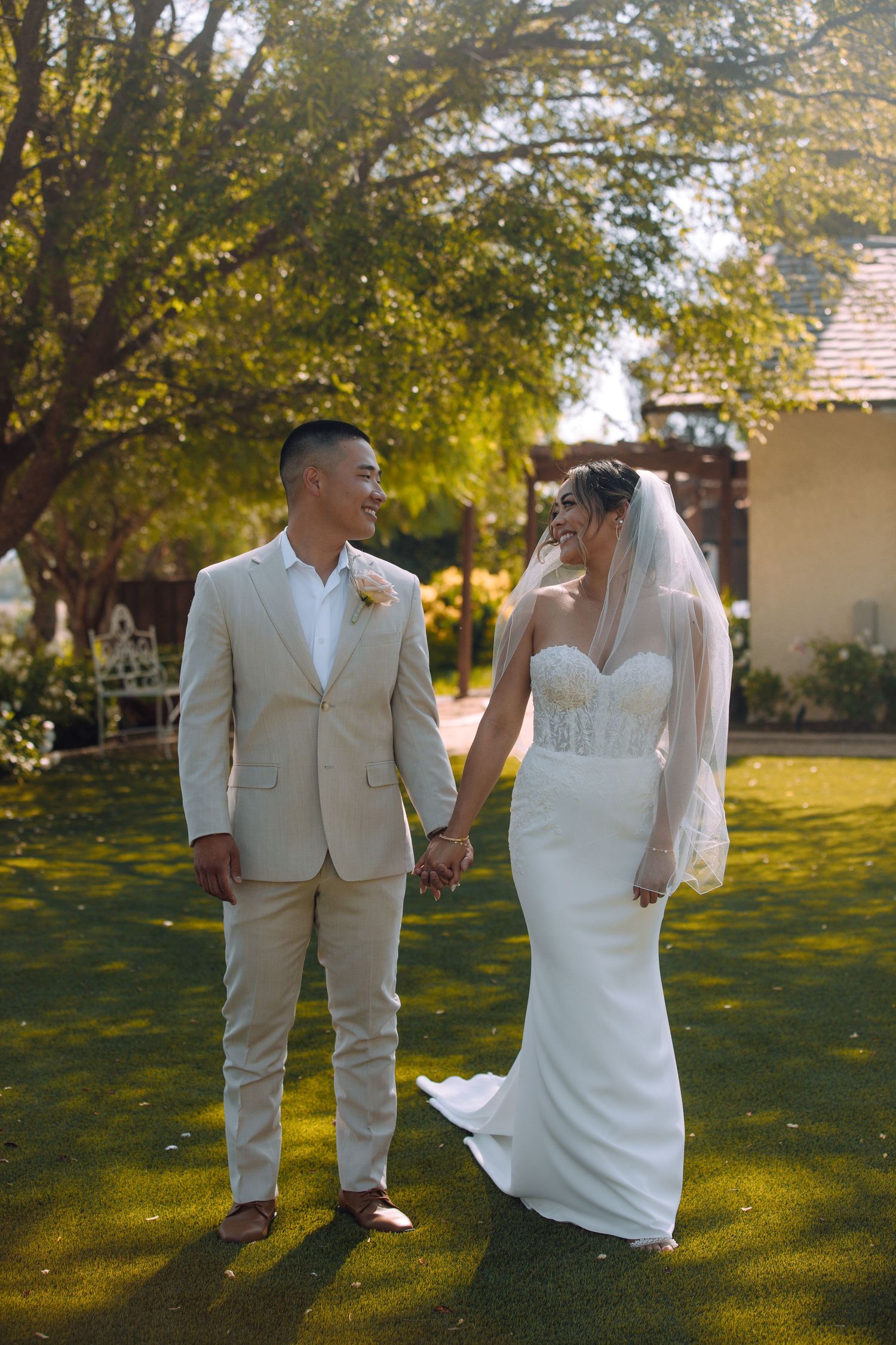 A couple in wedding attire stands together on a sunlit lawn.