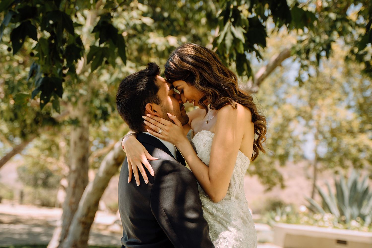 A romantic wedding couple embraces and kisses under sunlit trees in an outdoor garden setting.