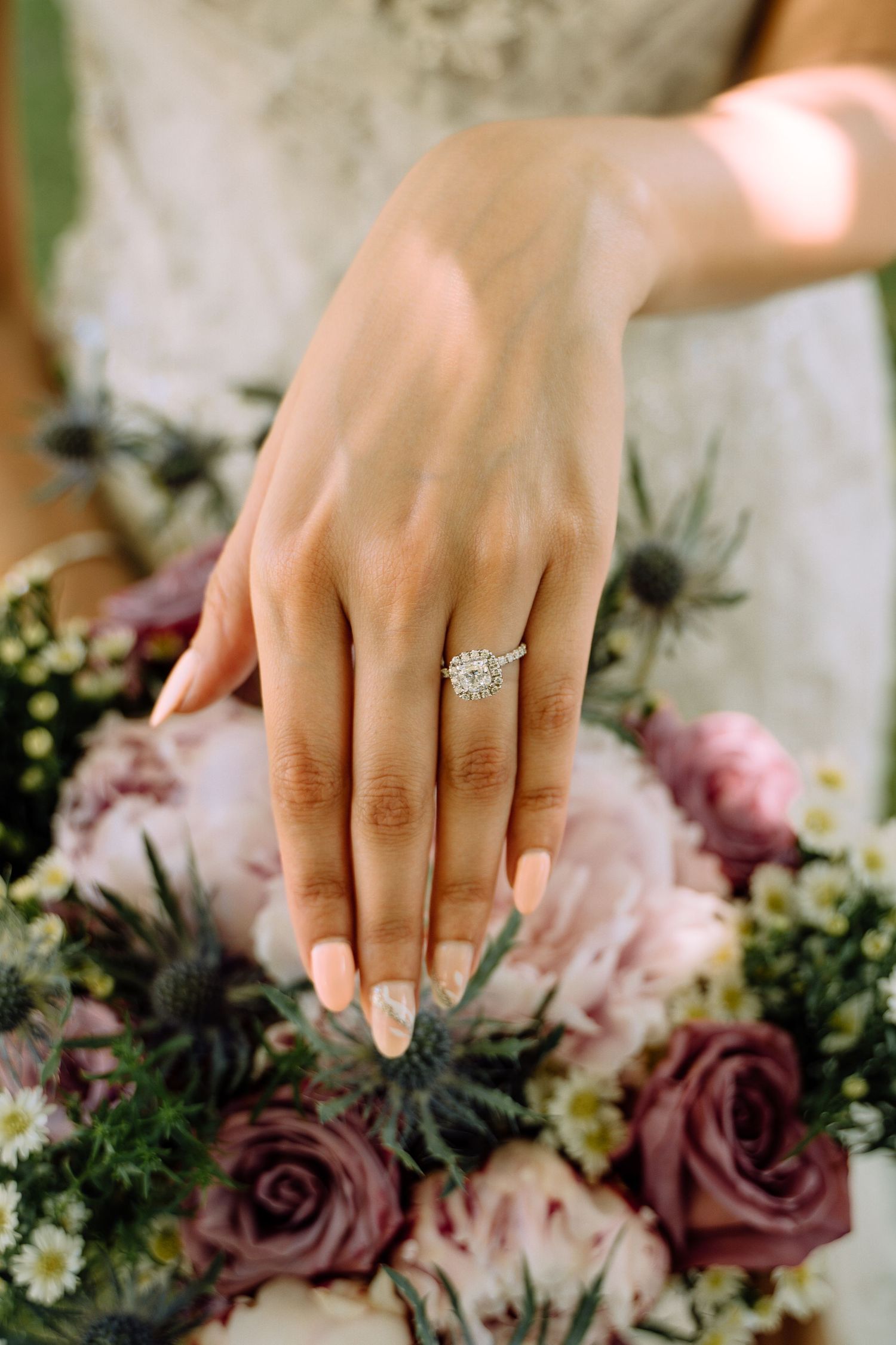 A delicate hand wearing a sparkling diamond engagement ring rests above a bouquet of pink and mauve roses.