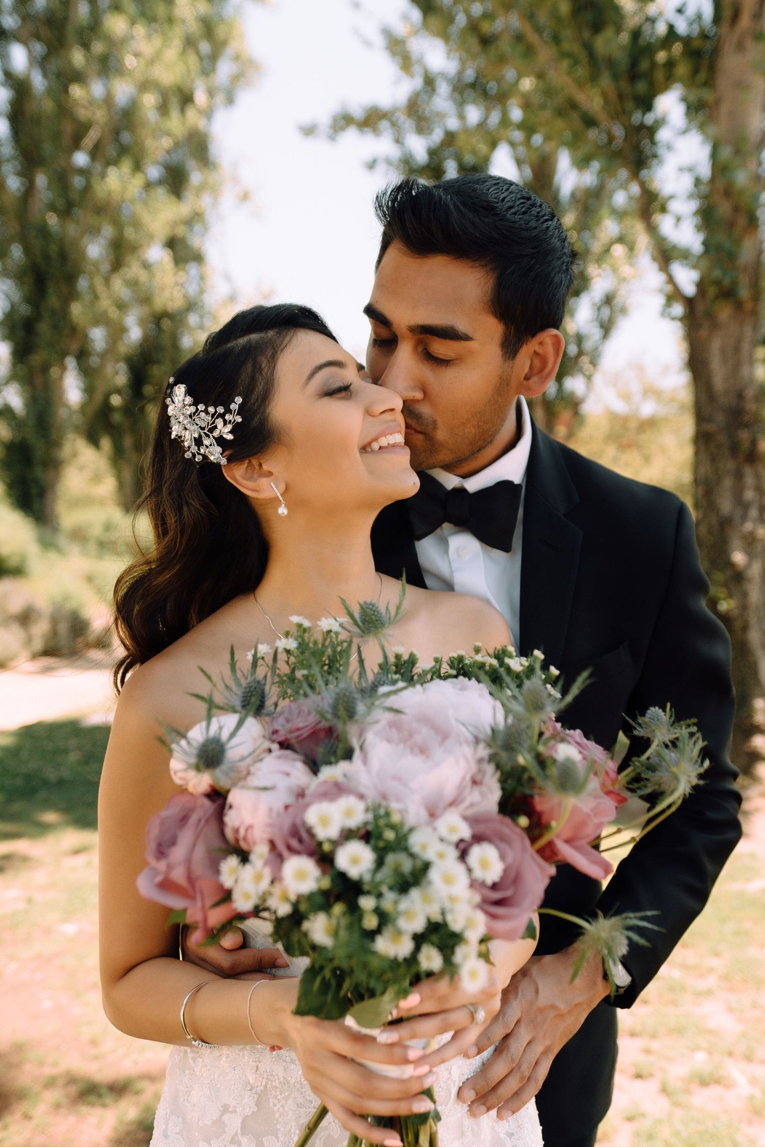 Bride holding pink peony bouquet while groom kisses her cheek in garden setting.