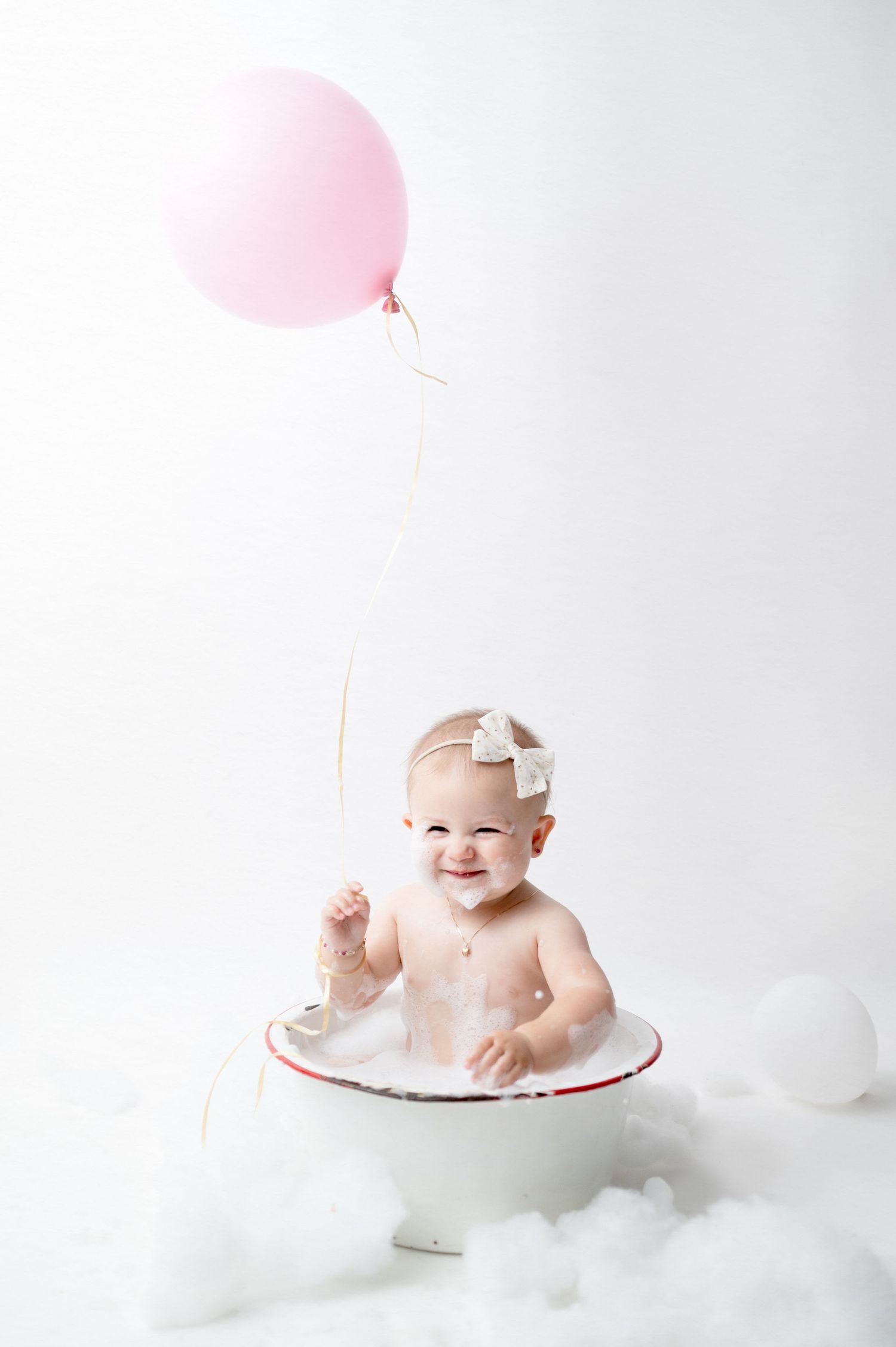 Joyful baby holds pink balloon while sitting in white enamel bowl for milestone photos.
