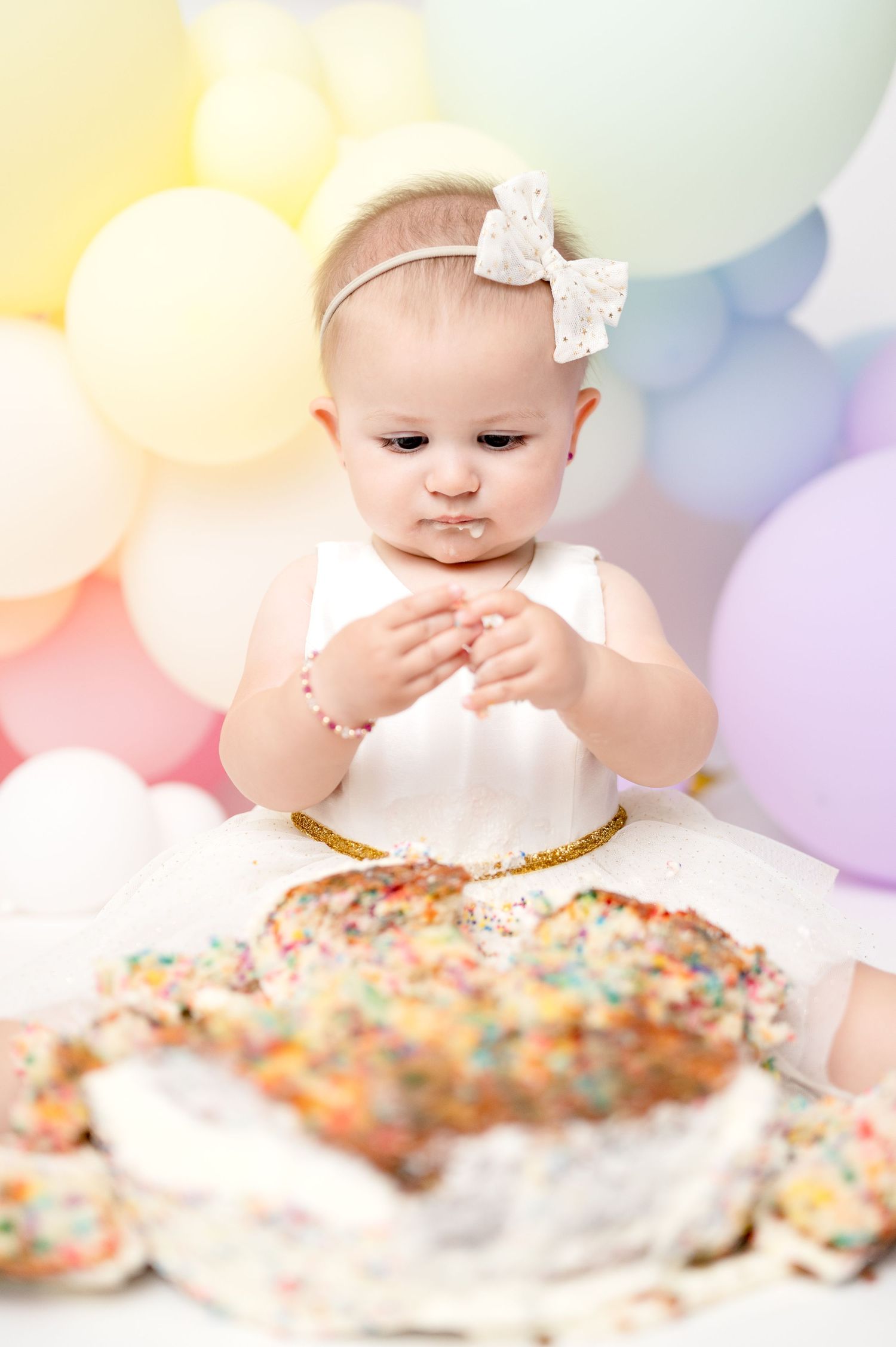 Baby in white dress examines sprinkled cake during first birthday cake smash photoshoot.