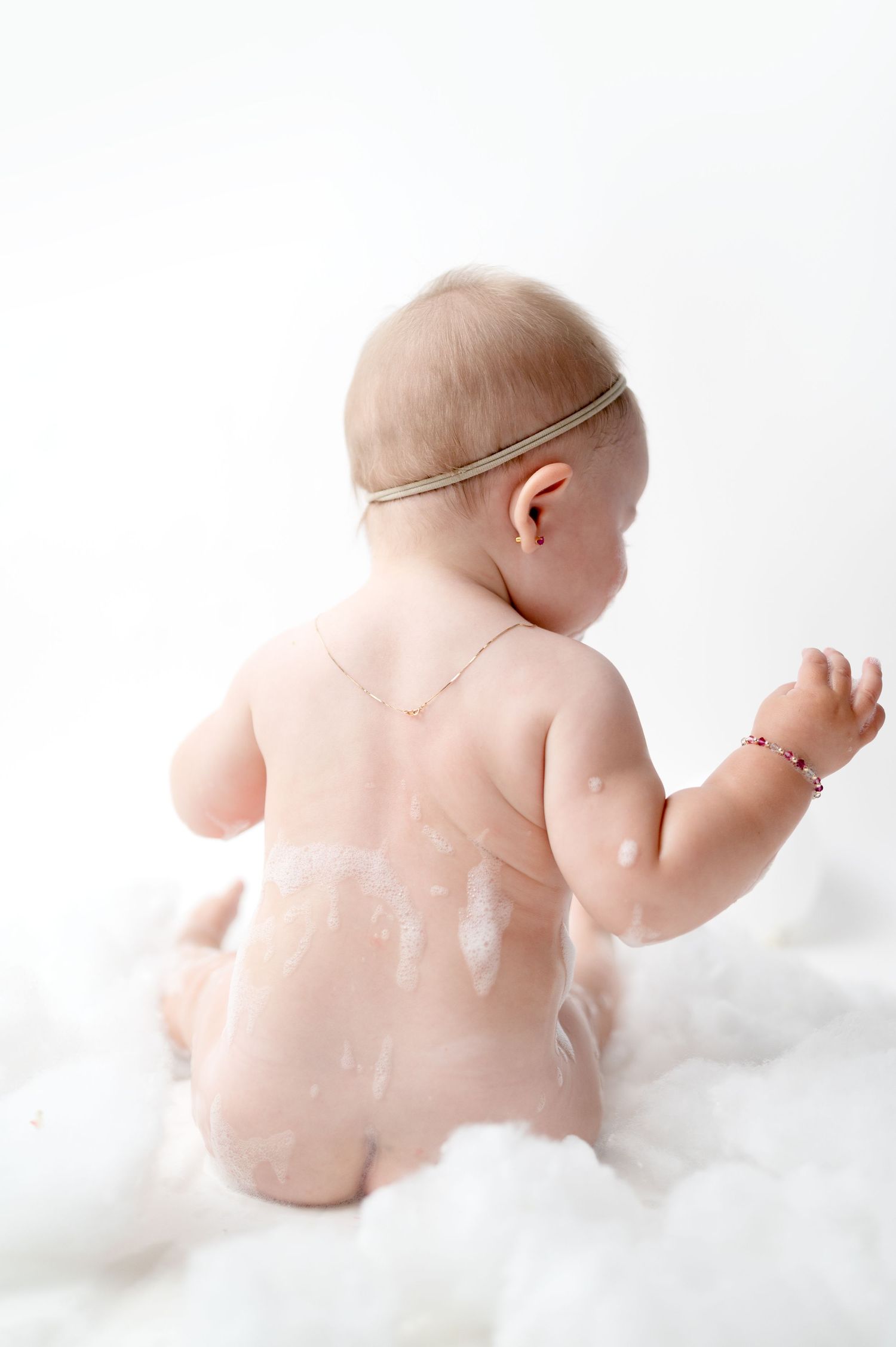 Baby wearing delicate headband sits among soft clouds in artistic studio portrait.
