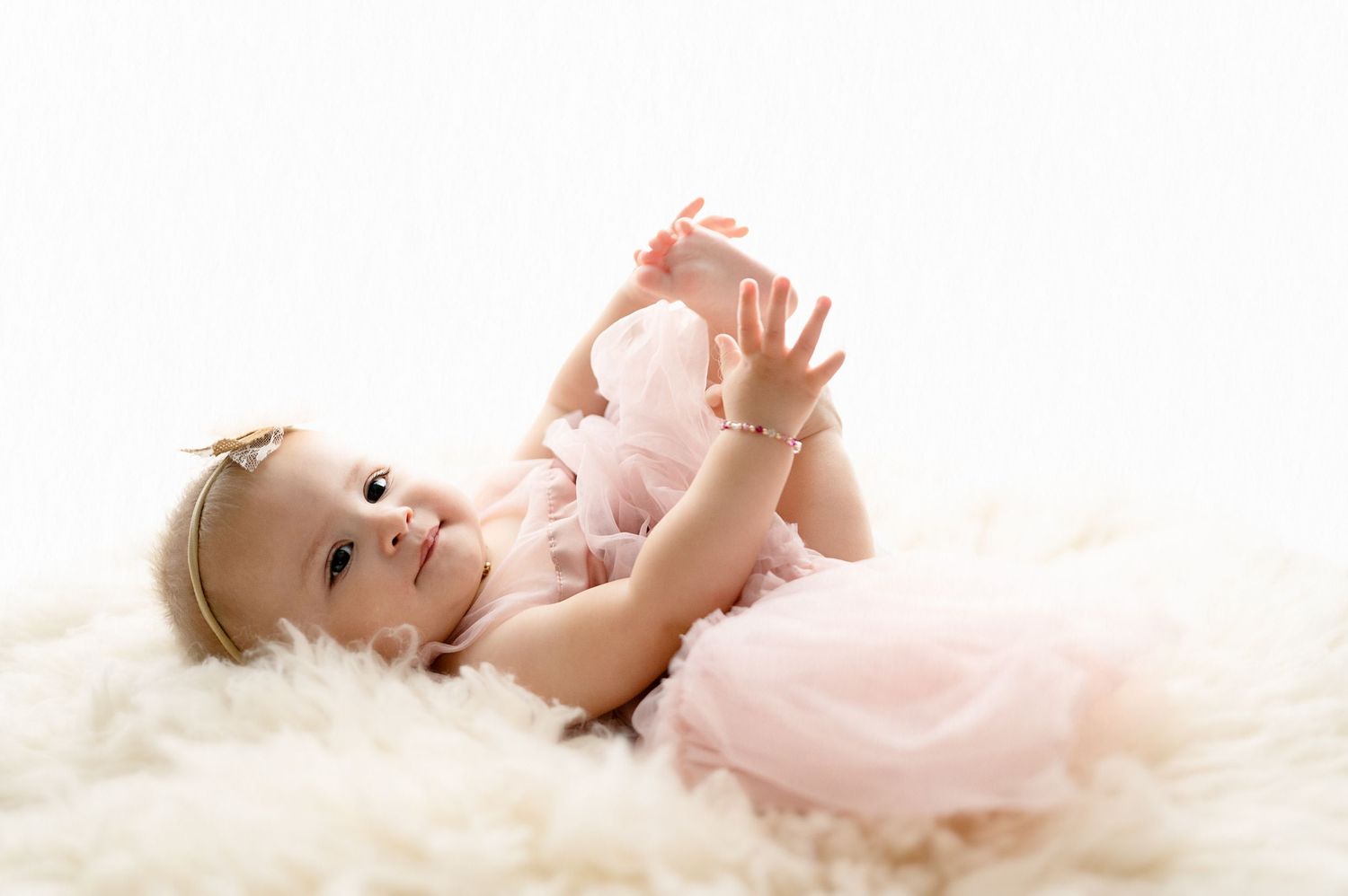 Baby in pink dress lies on soft white fur during professional photo session.