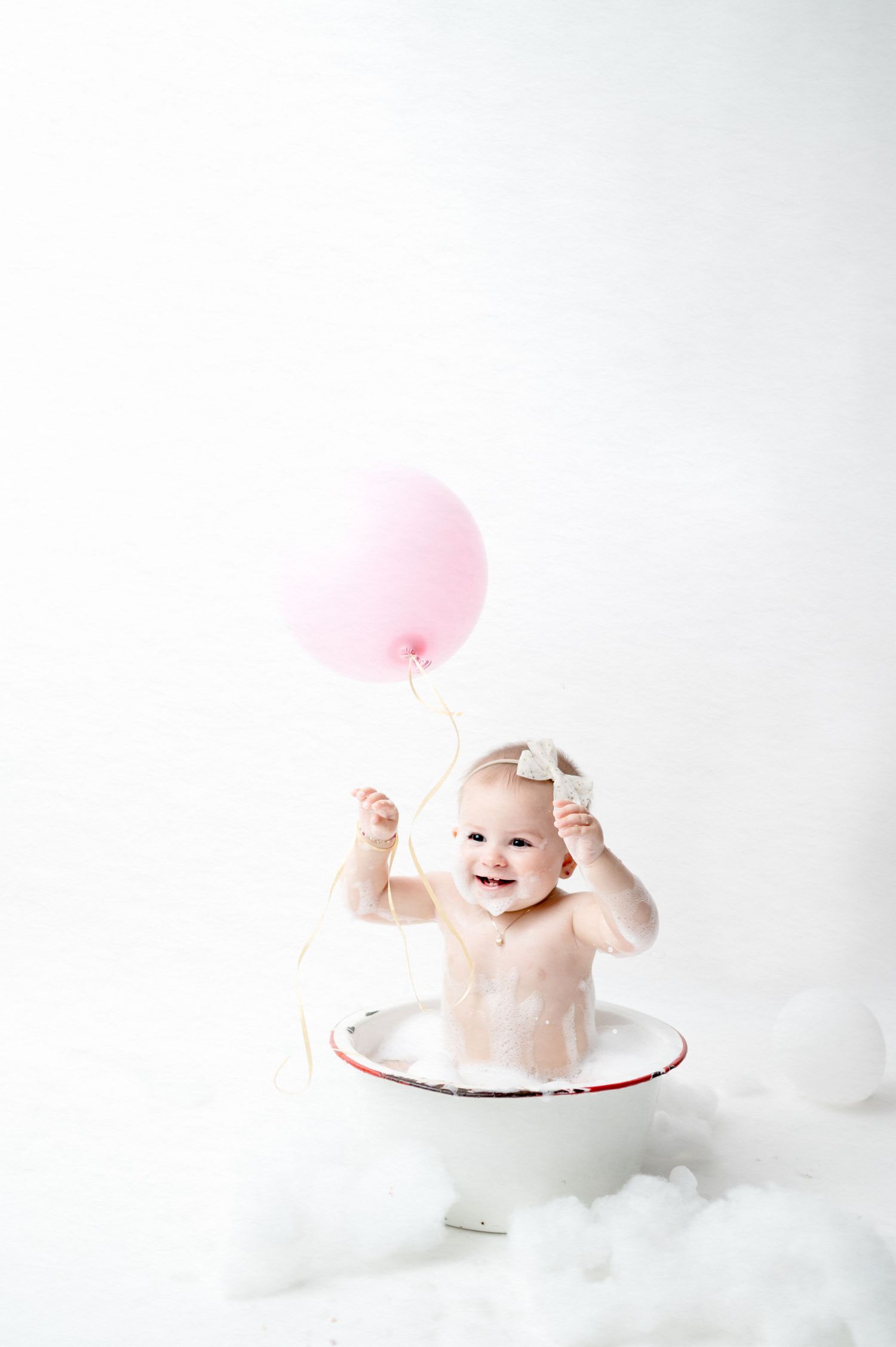Small child playing with pink balloon in white bathtub during minimalist photo session.