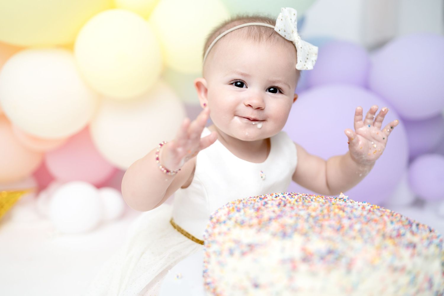 Baby wearing bow headband smiles near colorful balloons and sparkly cake during first birthday shoot.
