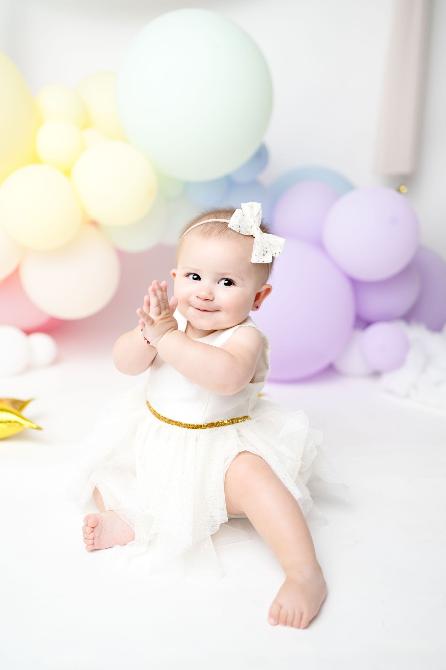 Baby in white dress smiles during celebration photoshoot with pastel balloon arch backdrop.