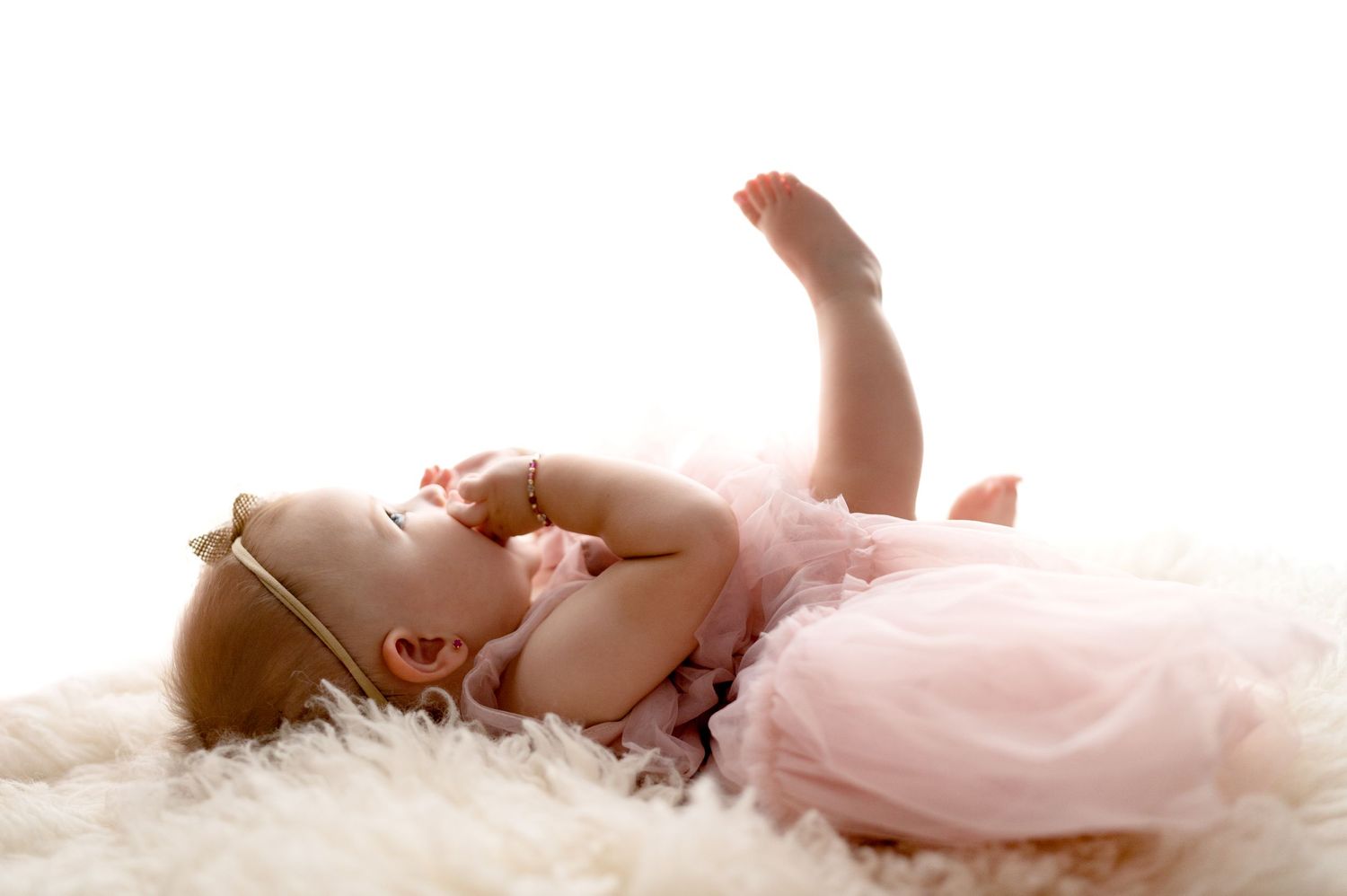 Baby in pink tulle dress lies on white fluffy rug in dreamy backlit portrait.