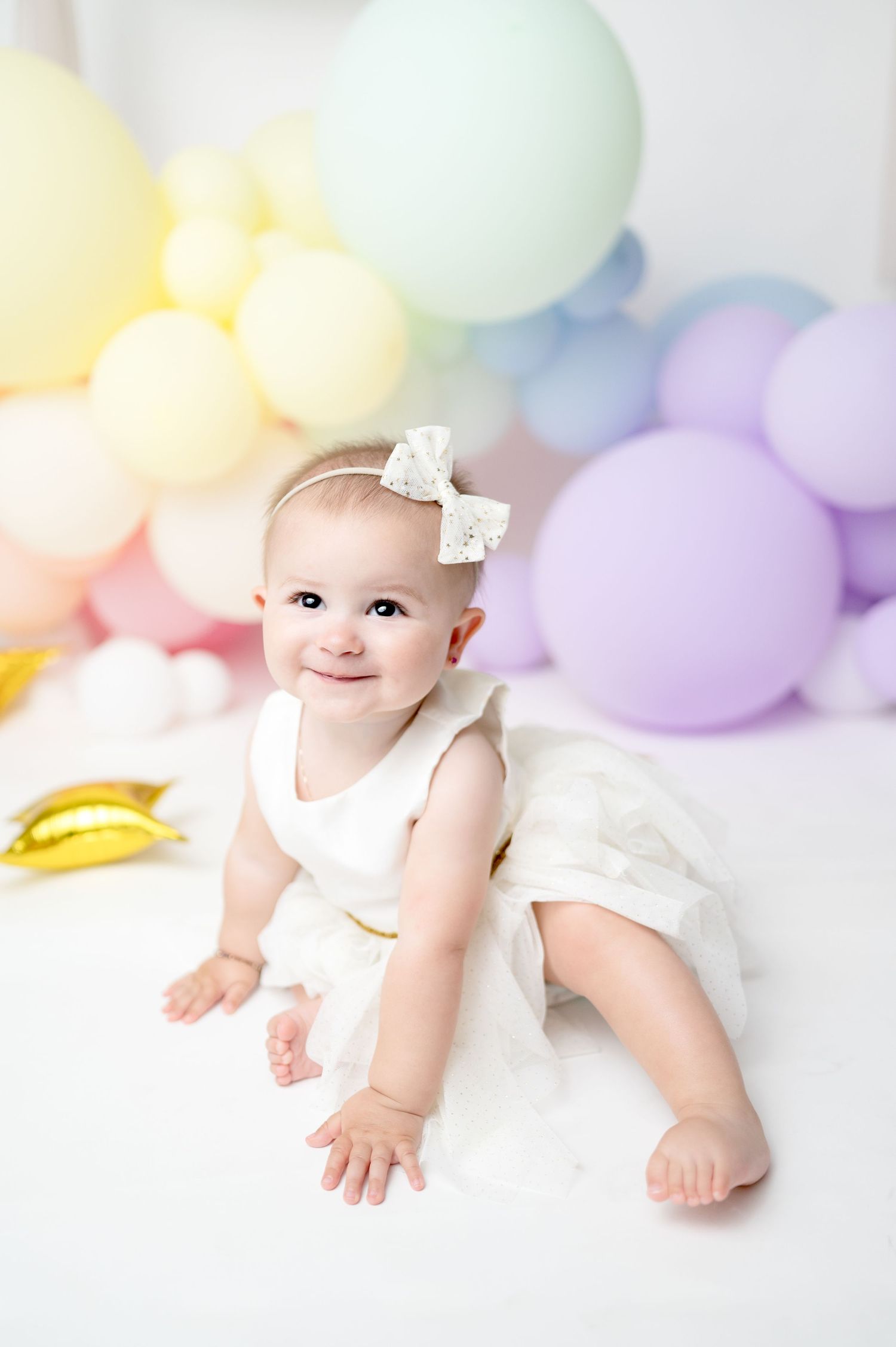 Baby in white dress smiles while sitting against pastel balloon backdrop for birthday photoshoot.