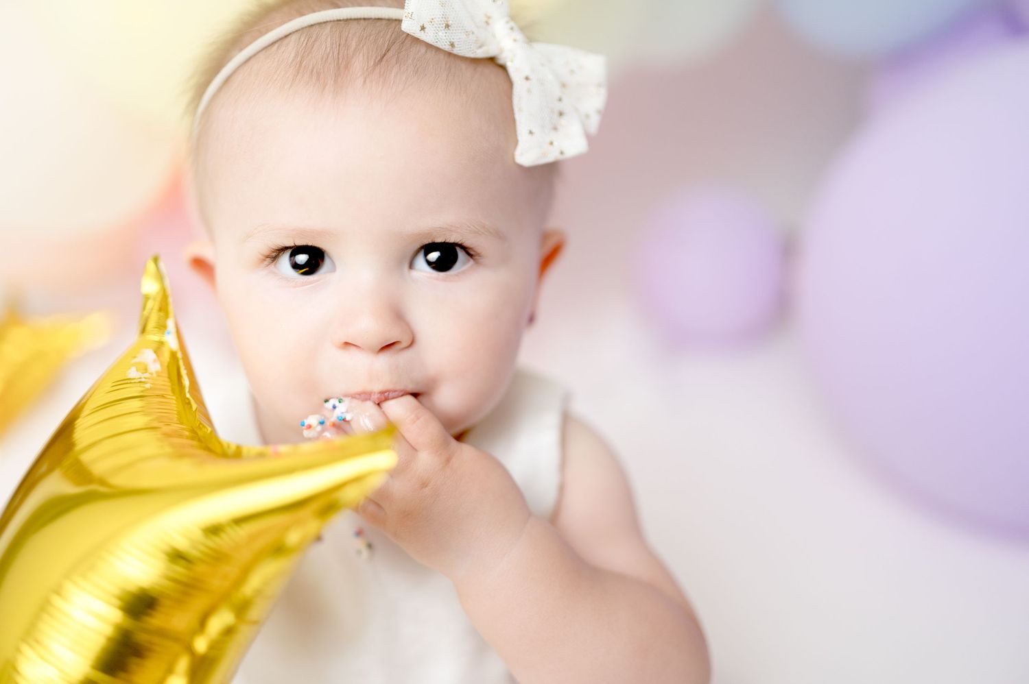 Baby wearing white bow headband nibbles on gold balloon during celebration photo session.