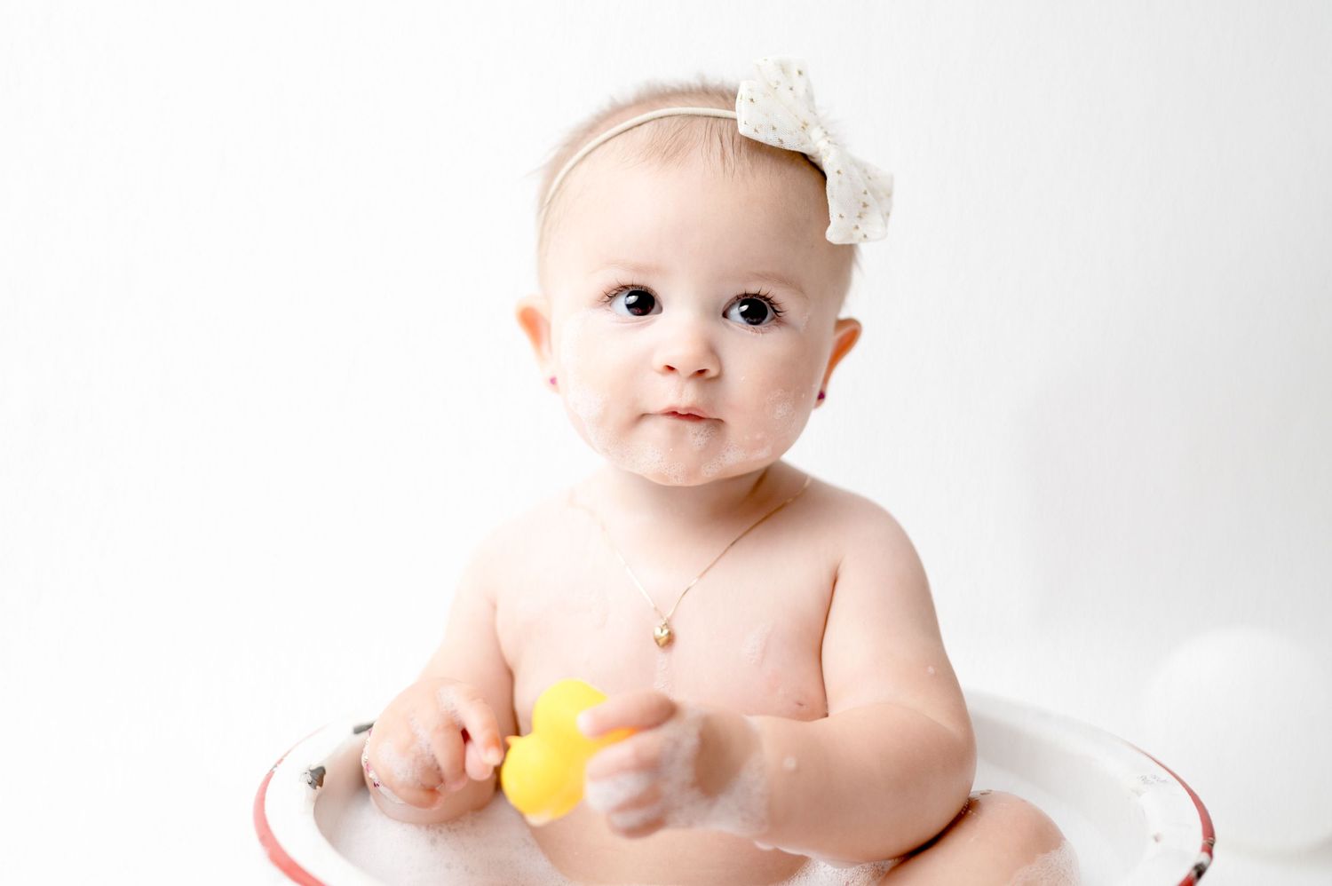Baby in white bow headband holds yellow toy during milestone photo session in vintage bowl.