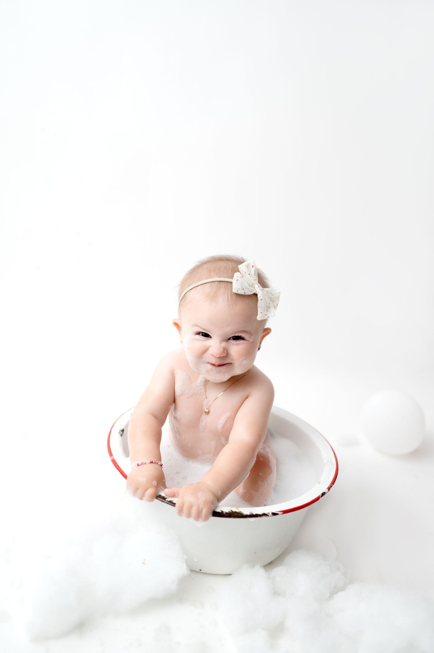 Smiling baby poses in rustic white enamel bowl surrounded by soft white decorations.