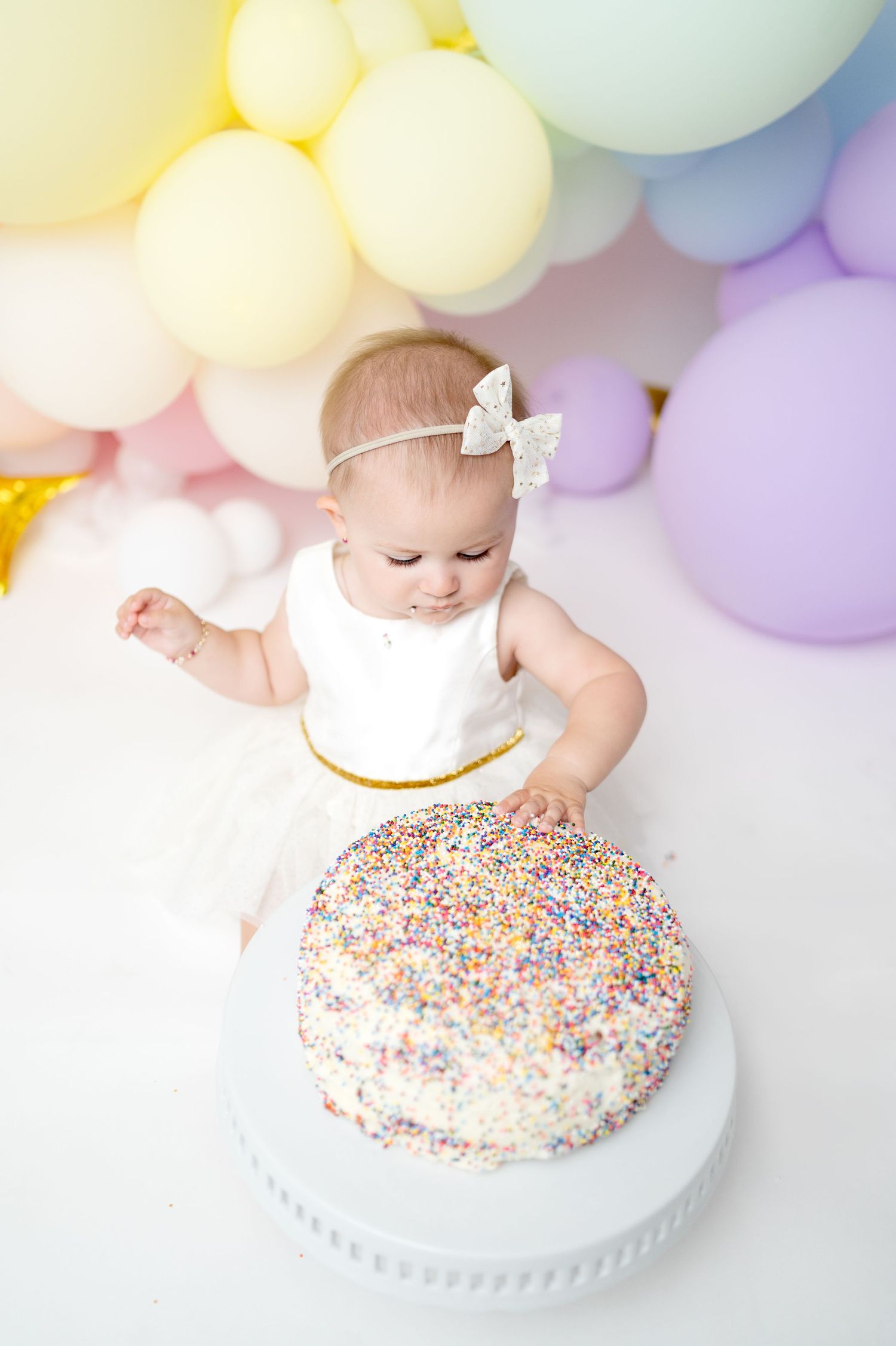Baby in white outfit reaches for sprinkle-covered birthday cake with pastel balloons in background.