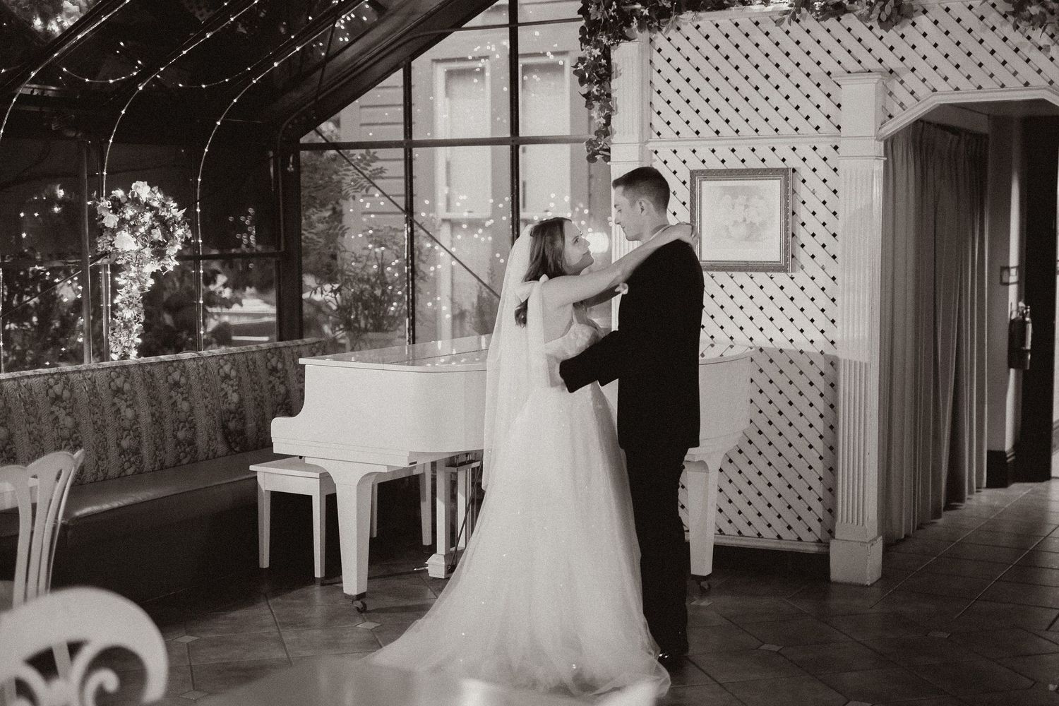 Black and white photo of couple dancing near white piano in an intimate wedding venue.