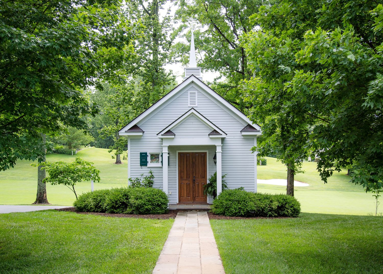 Small white chapel with wooden door surrounded by lush green trees on sunny day.