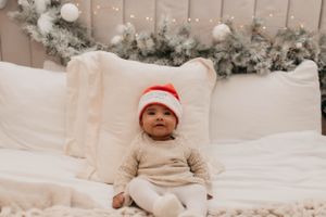 Baby wearing Santa hat lying on white bedding with holiday decorations.