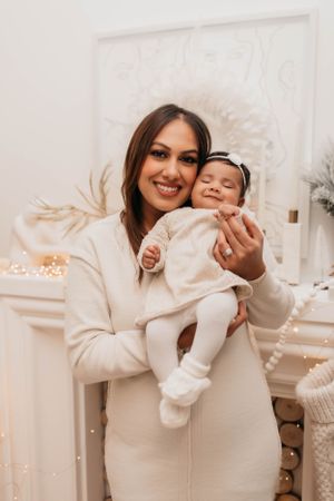 Joyful holiday portrait in cream tones with parent and baby against festive backdrop with lights.