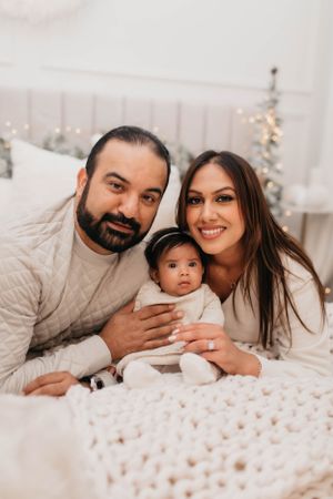 A joyful family of three poses together on a white bed with cozy knit blanket and holiday decor.