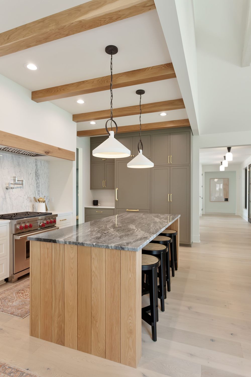 Modern kitchen featuring wood and gray cabinetry, marble island, and white pendant lights with exposed wooden ceiling beams.