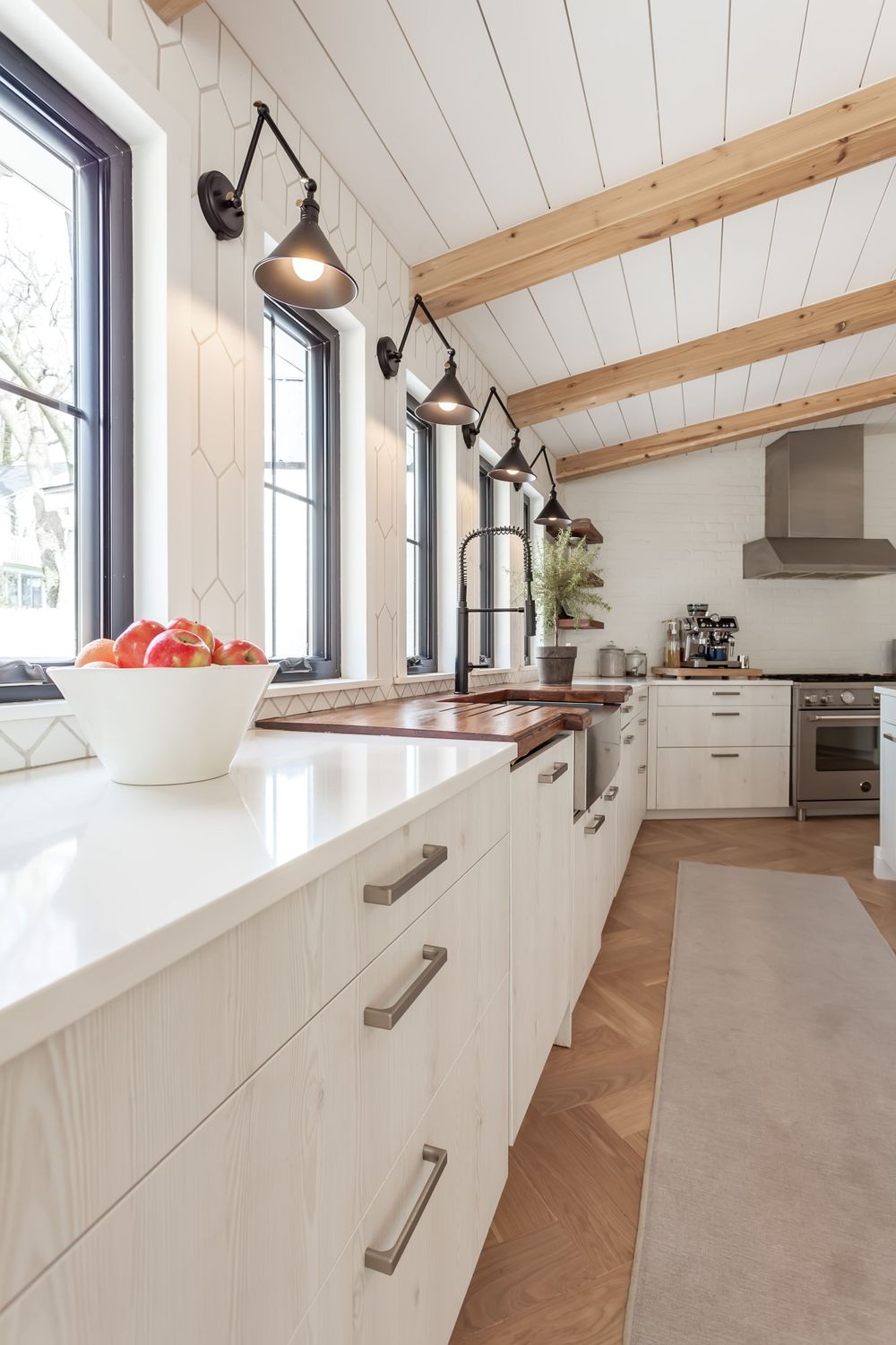 Modern white kitchen with wood beams, black sconces, and sleek cabinet design.