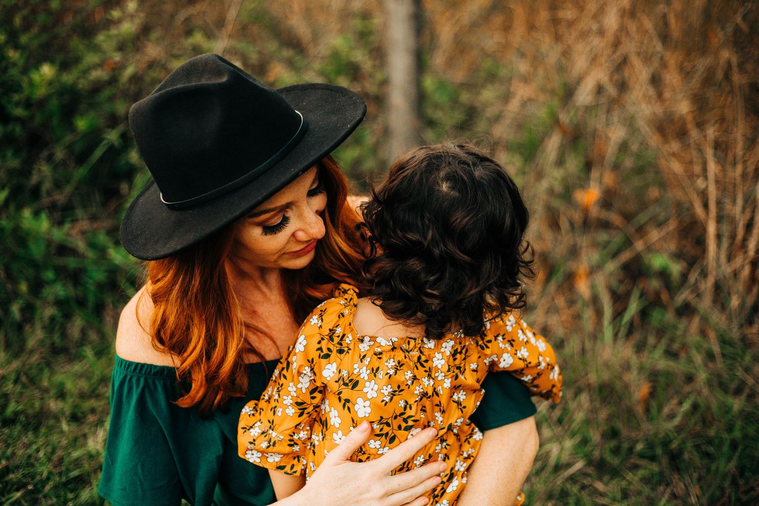 Mother in black hat holding young child wearing yellow floral dress against natural outdoor backdrop at sunset.