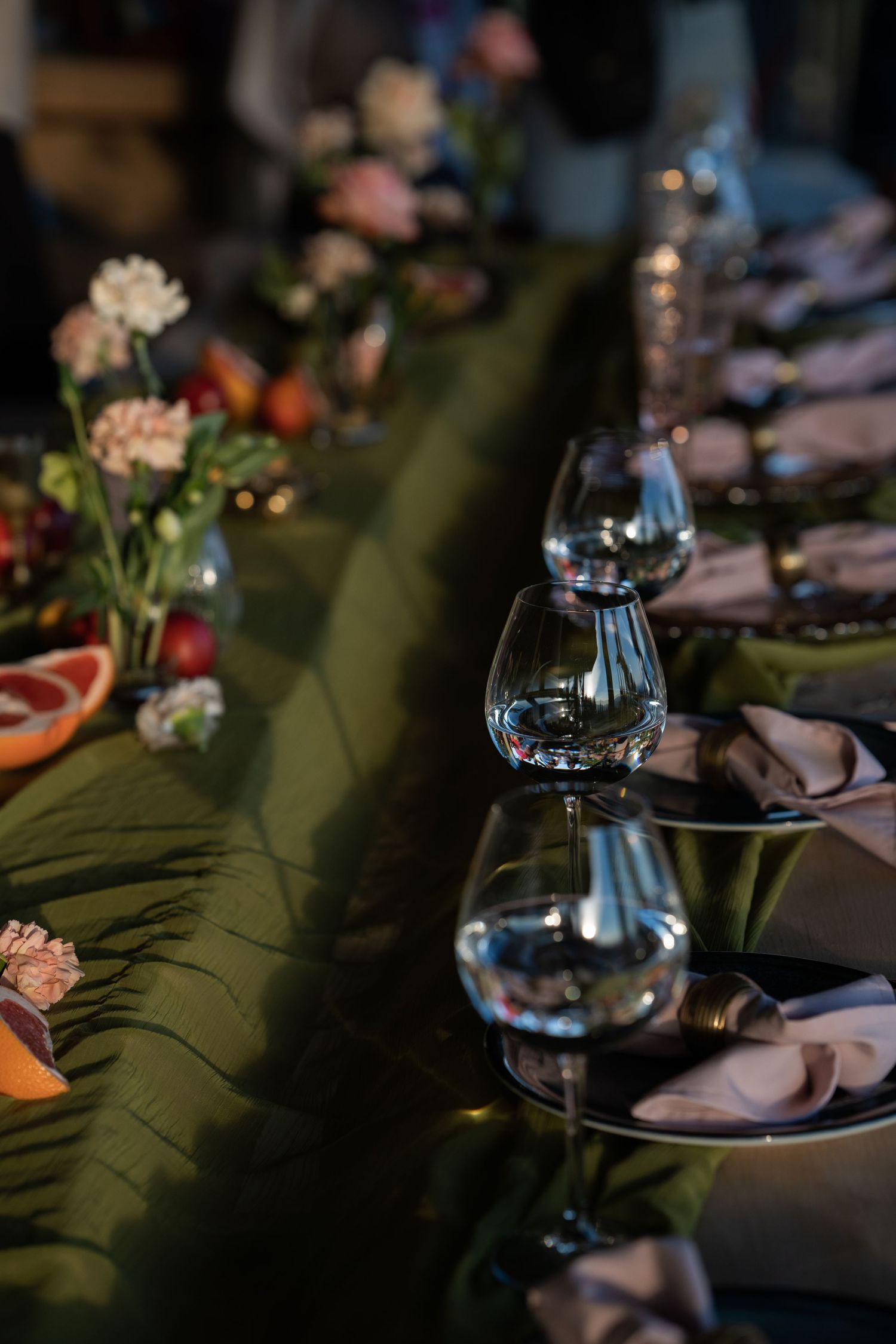 Long dining table with green runner, wine glasses, pink napkins, and floral centerpieces.