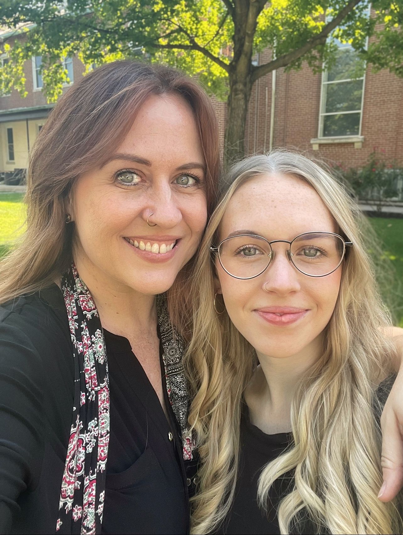 Outdoor selfie on a sunny day with brick building and green trees in background.