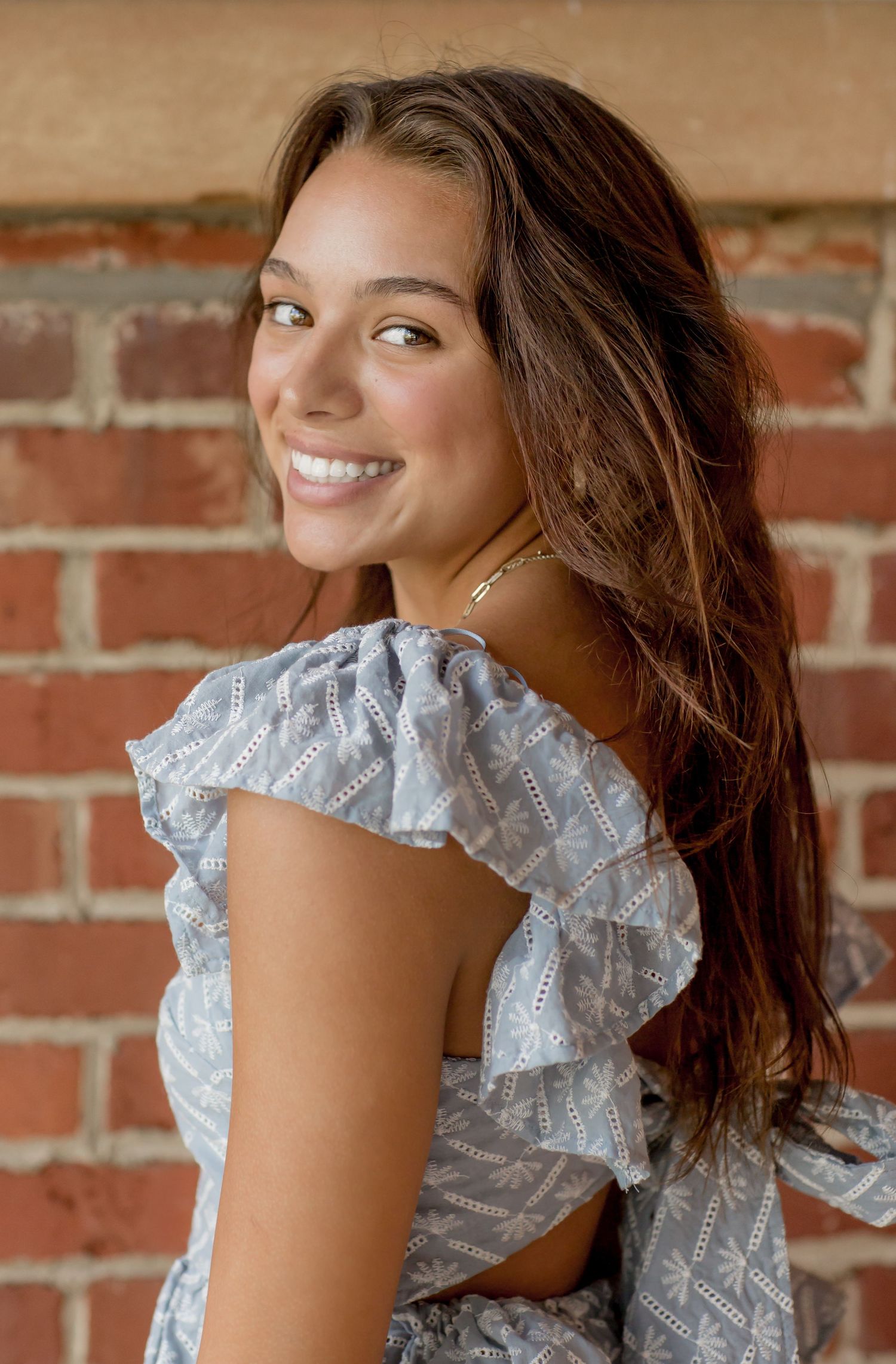 Young woman in ruffled blue top smiling over shoulder against brick wall.