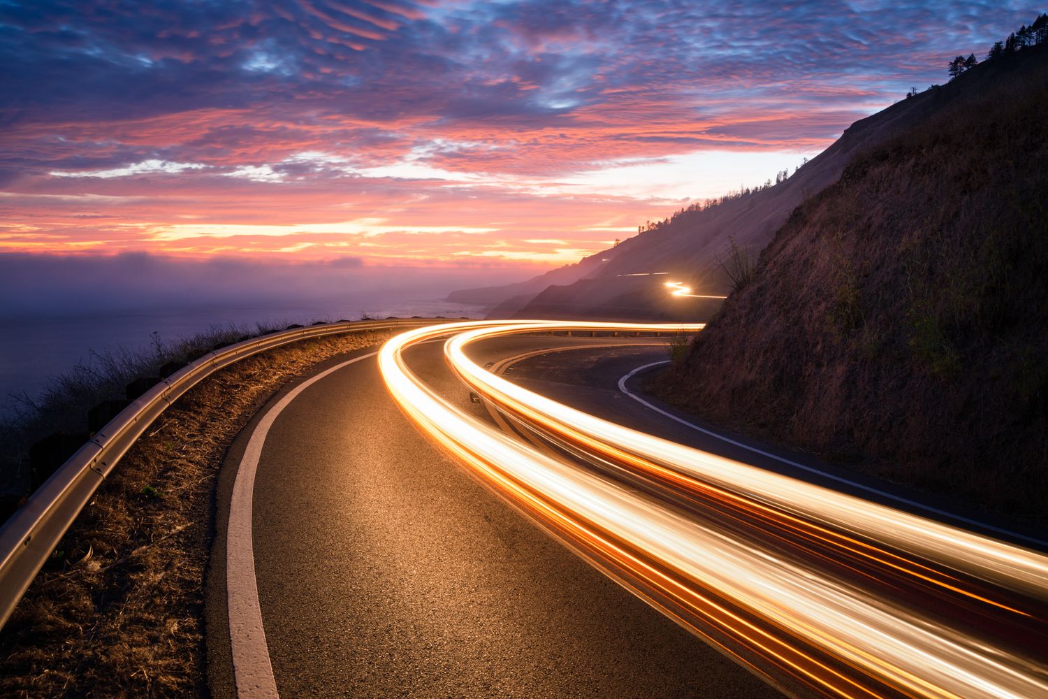 Long exposure of car light trails along curved coastal highway at dramatic sunset.