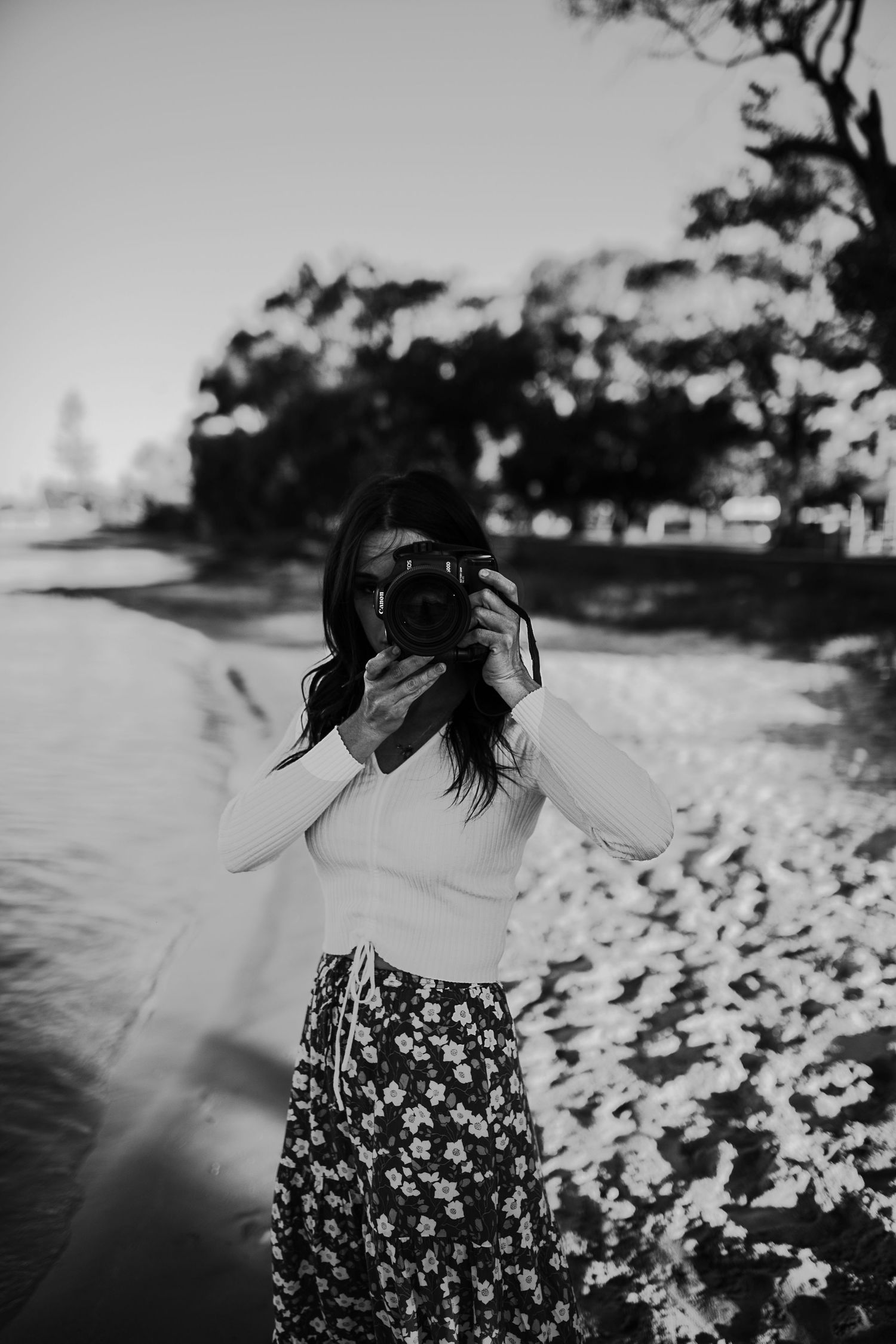 A photographer takes pictures in a flowing floral dress by the water at sunset.