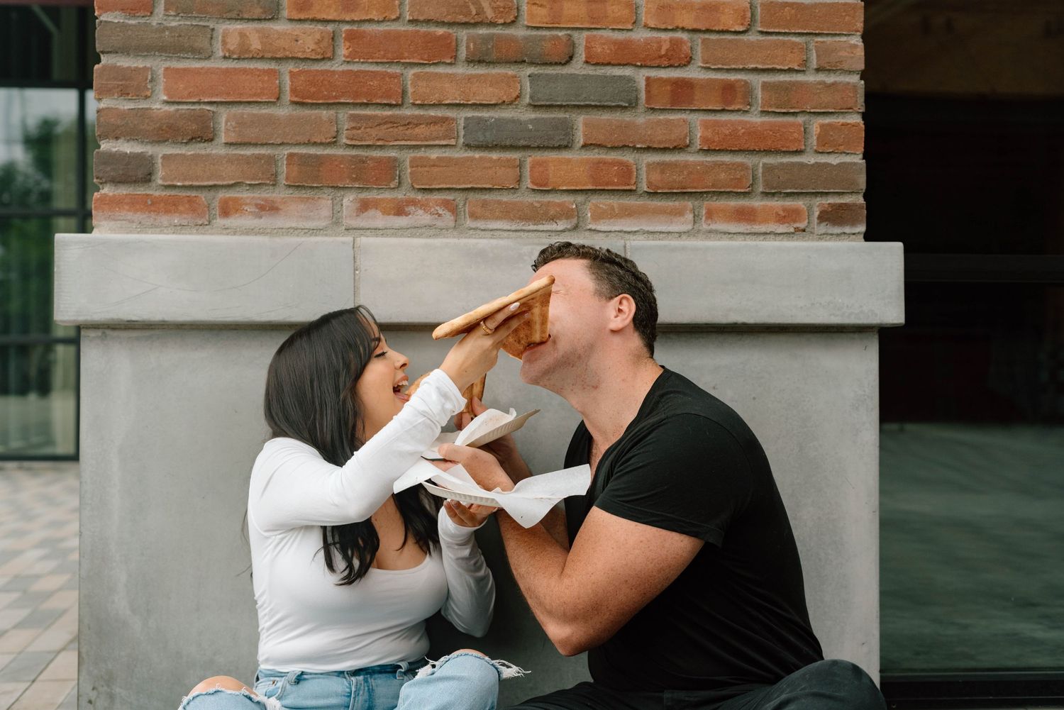 A couple poses while eating a slice of pizza in an urban setting during a photography session in Los Angeles, California