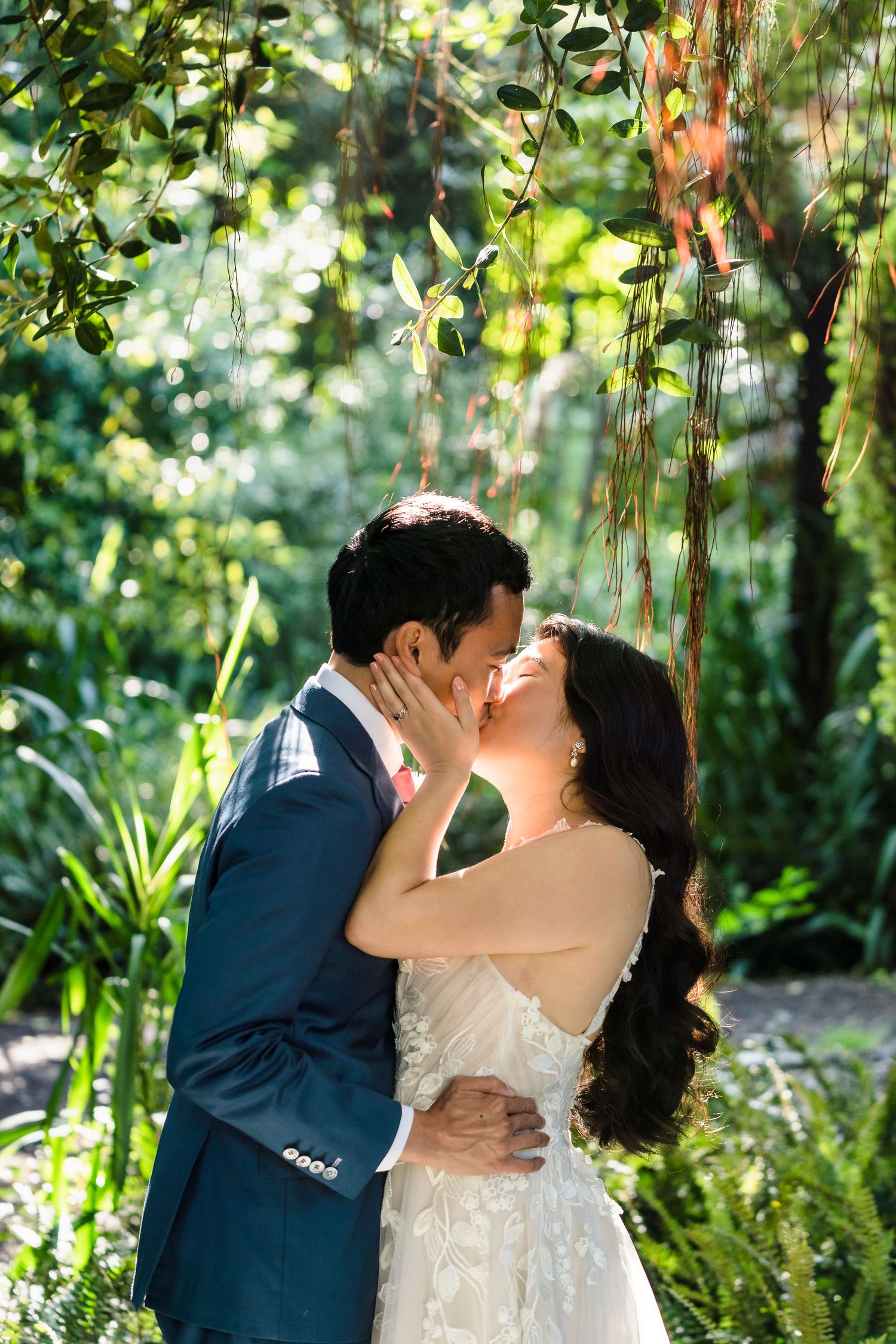 A romantic wedding couple shares an intimate kiss under hanging vines in a lush garden setting.