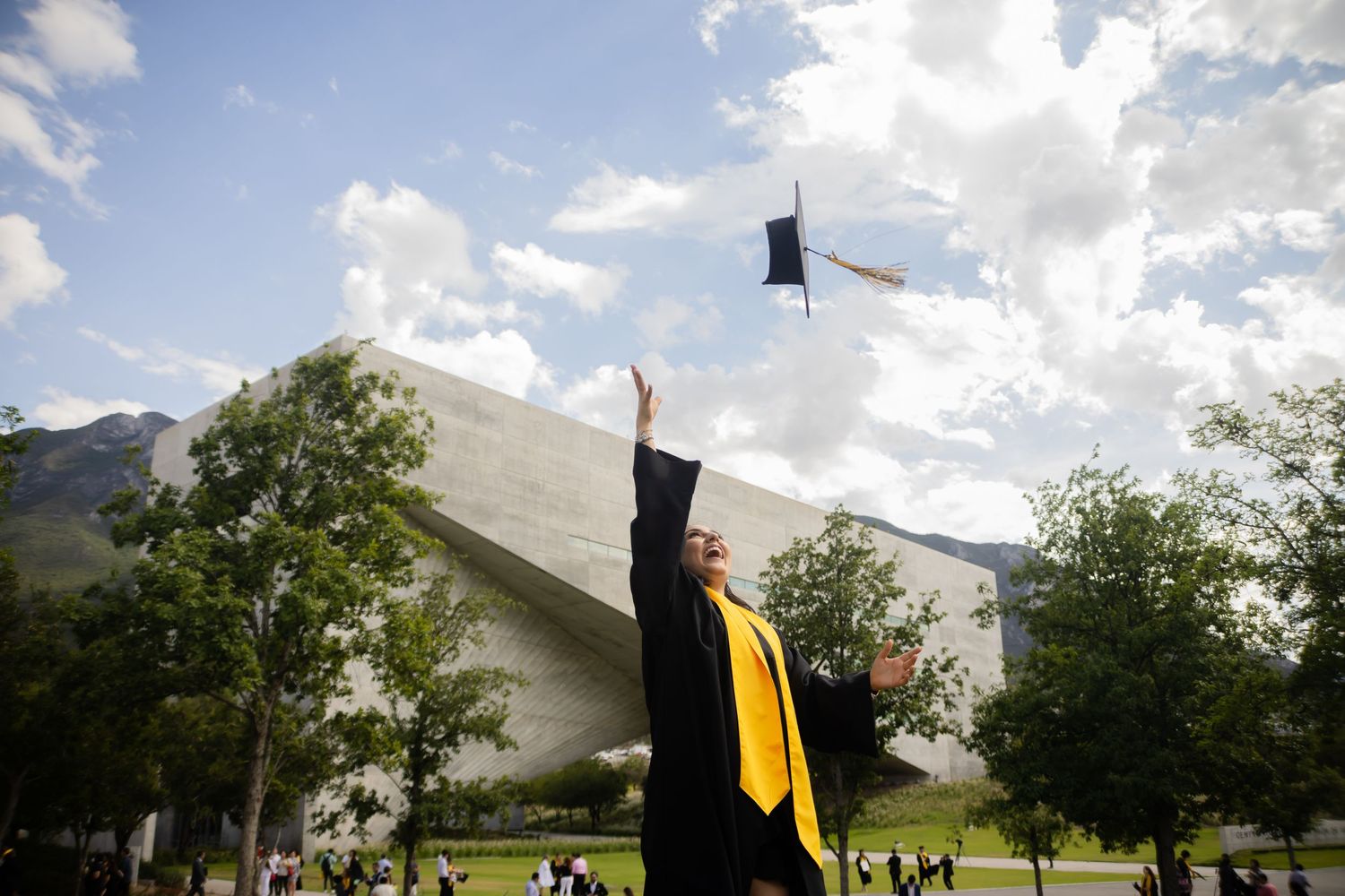 Graduate in black and gold gown tossing mortarboard cap into blue sky outdoors.
