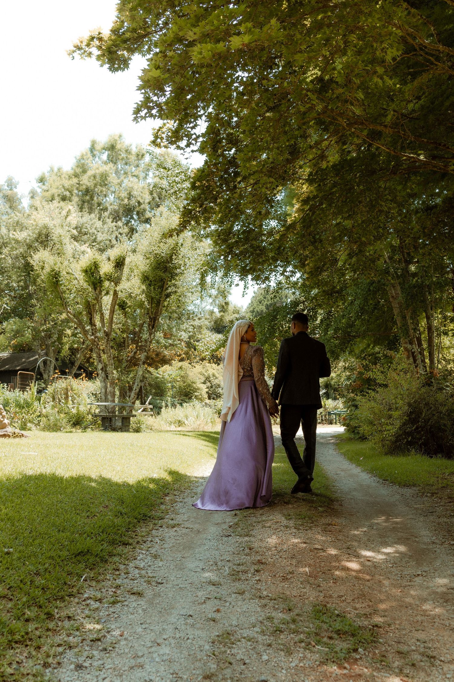 A couple walks down a tree-lined path, with the person in a flowing purple dress and another in black.