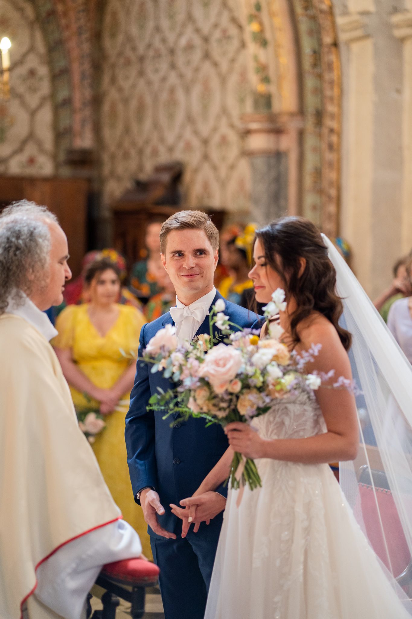 A wedding ceremony takes place in an ornate church with decorative walls and a priest officiating the service.