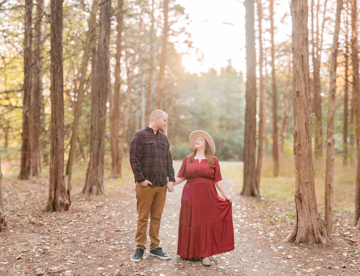 Couple holds hands walking through sunlit pine forest in golden autumn light wearing red dress.