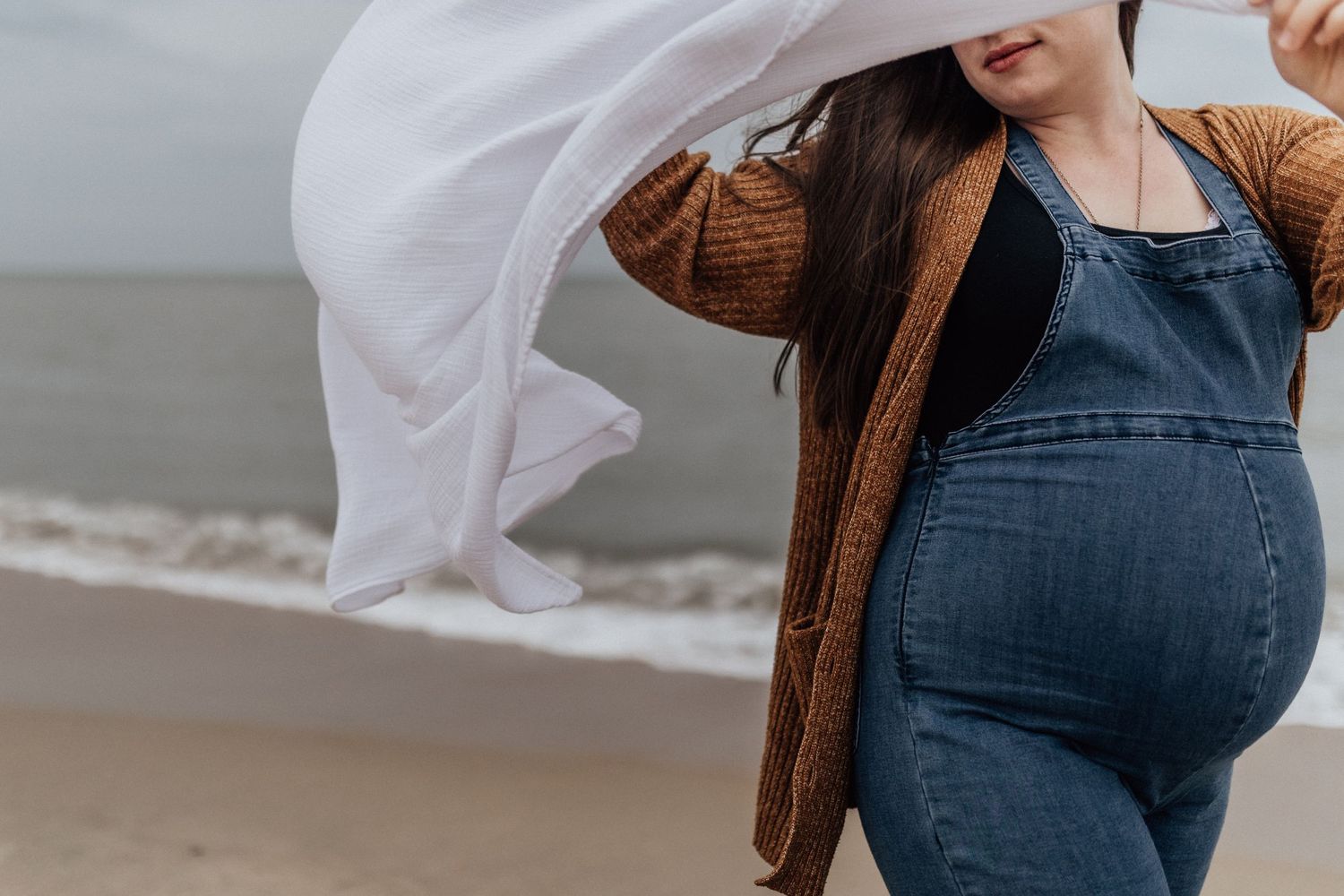 pregnant woman in overalls on beach in virginia beach with white baby blanket