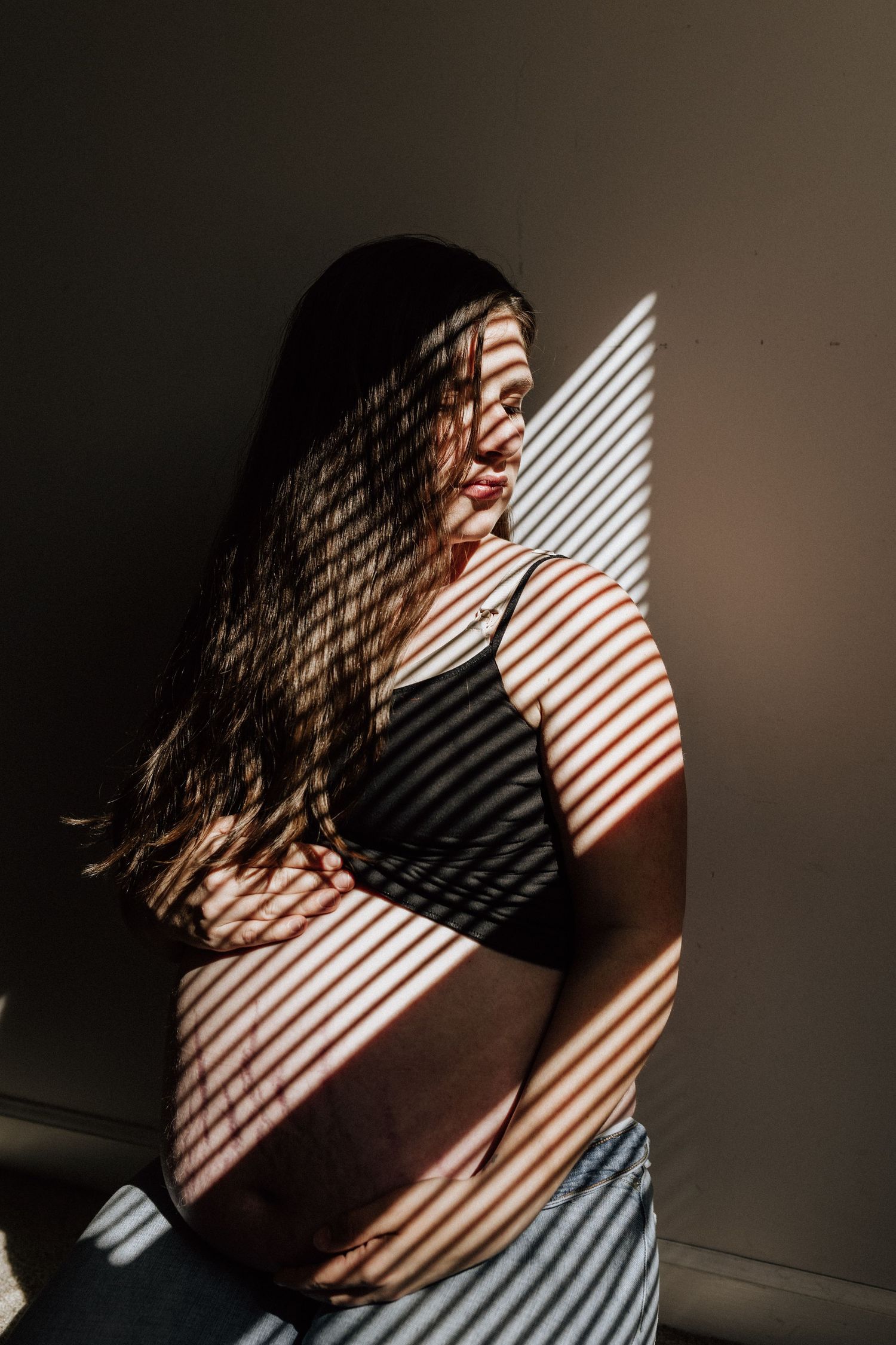 pregnant woman sitting in the window light and shadows of the blinds in her home