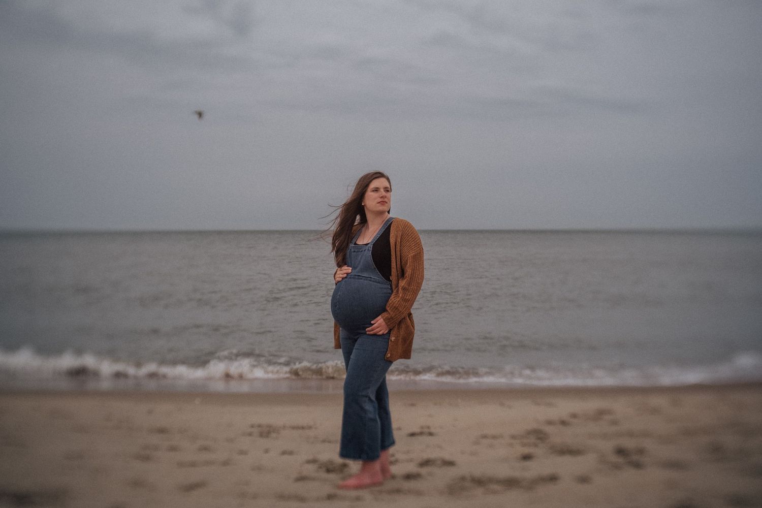 pregnant woman on the beach in virginia beach wearing overalls and cardigan