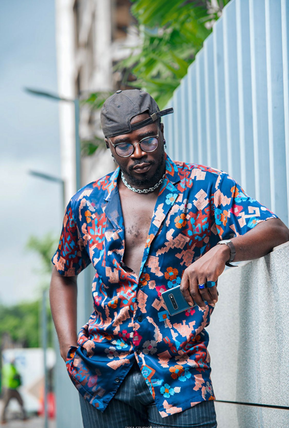 Person in colorful patterned shirt and glasses poses casually against white pillars outdoors.