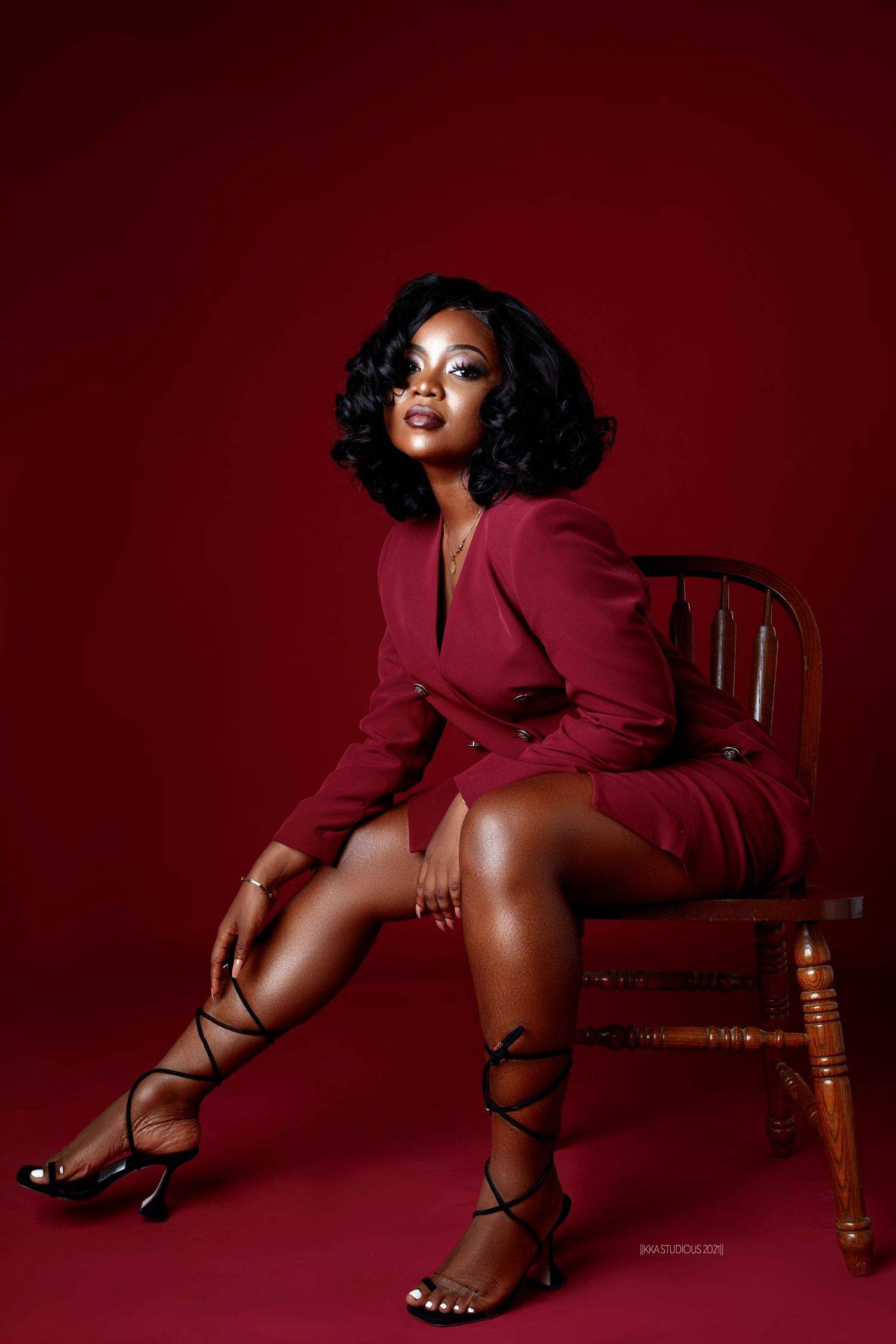 Fashion model poses dramatically against deep red backdrop wearing burgundy blazer and strappy heels.