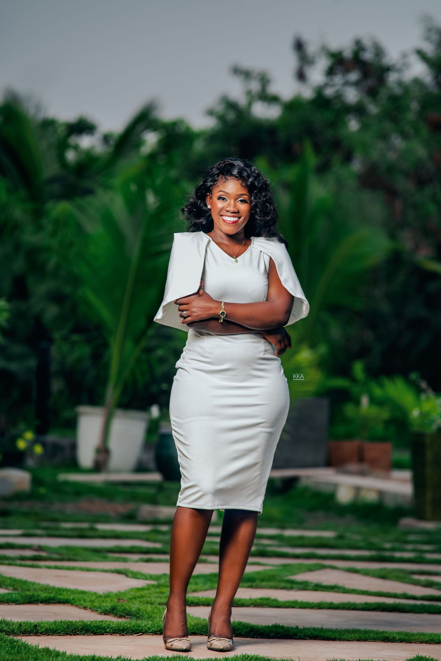 Woman in white dress poses joyfully outdoors in a lush garden setting with stone pavers.