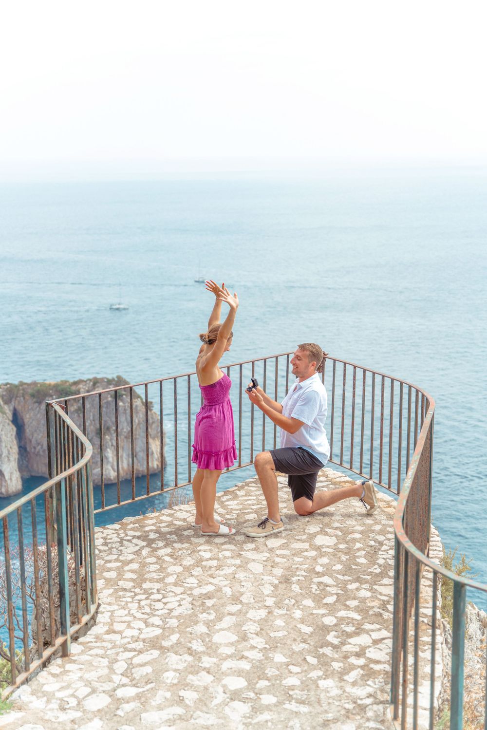 Couple posing playfully on stone terrace overlooking Mediterranean Sea.