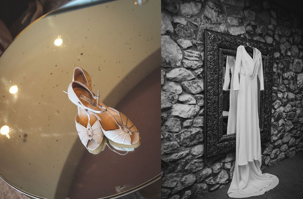 Wedding details featuring white bridal shoes and dress hanging against stone wall.