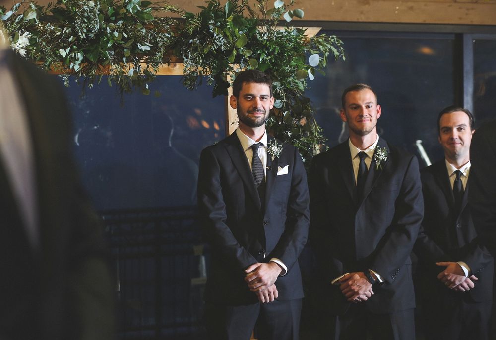 Wedding party members in dark suits standing in front of blue backdrop with greenery.