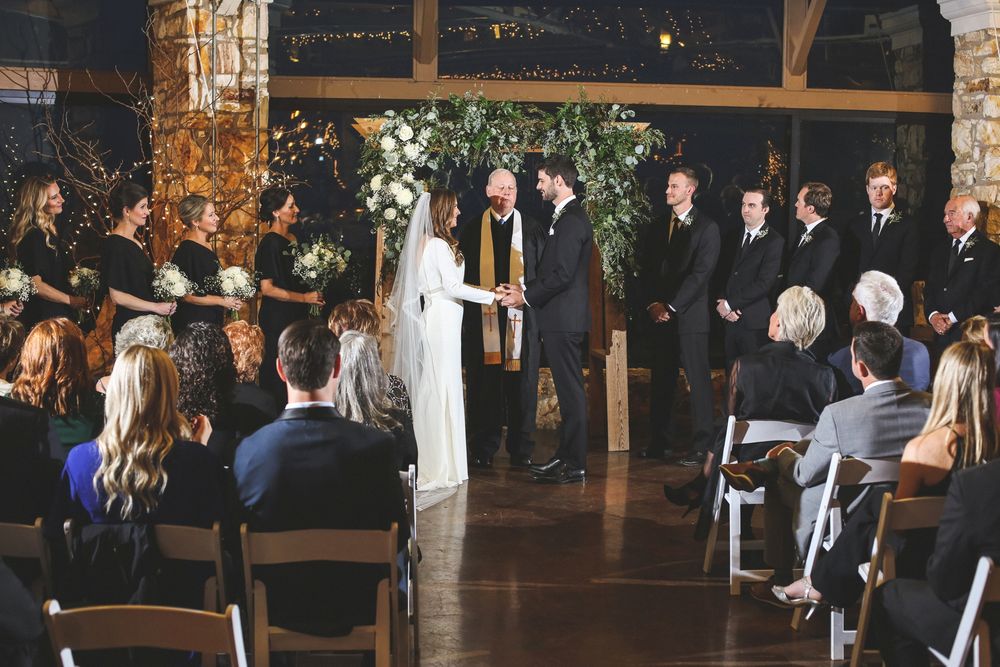Indoor wedding ceremony with guests seated in chairs and floral decorations overhead.