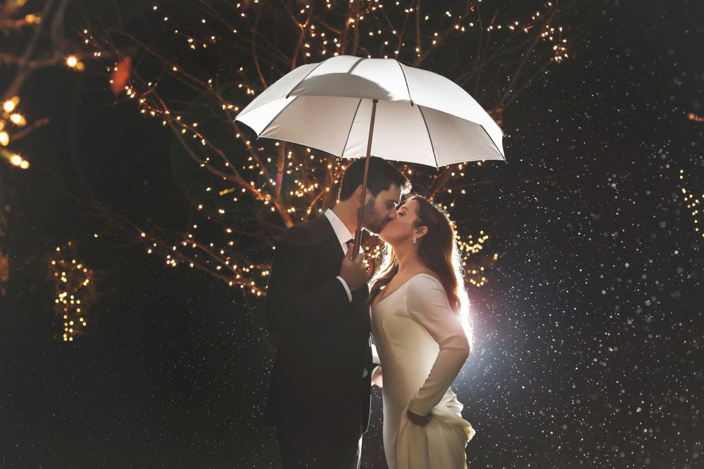 A romantic couple shares a kiss under a white umbrella surrounded by sparkling lights at night.