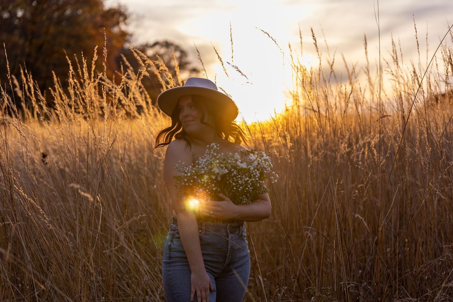 Woman in hat holds wildflower bouquet standing in golden tall grass field during stunning sunset.